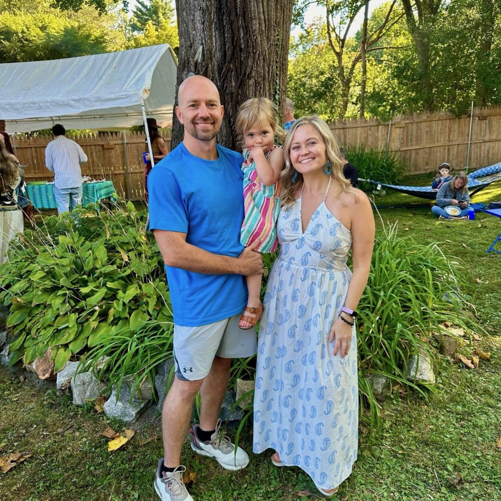 man in blue shirt smiling, holding a young girl and standing next to a smiling woman